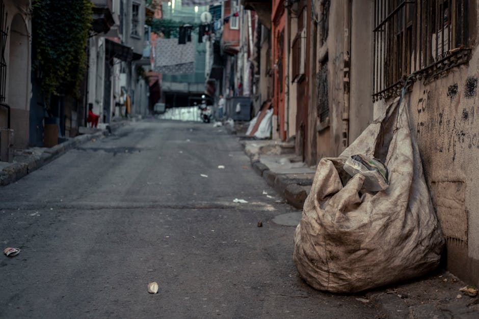 A collection of overflowing rubbish and waste materials is piled on a paved urban sidewalk, primarily consisting of cardboard boxes, paper bags, black and red plastic rubbish bins, and miscellaneous p