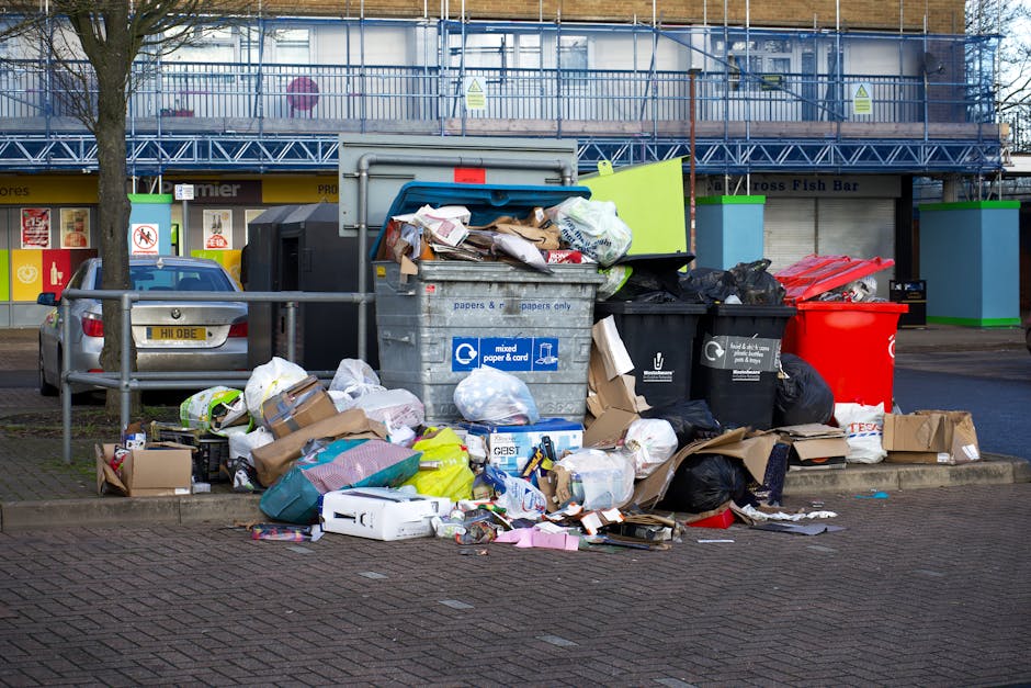 A street scene featuring a white garbage truck operated by a waste management company, with visible rust and grime on its rear compactor section. The truck is positioned close to the curb on a cobbles